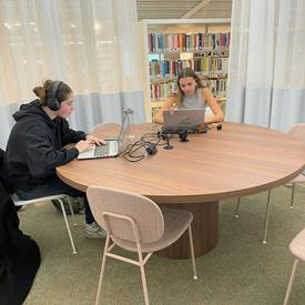 foto: Twee jonge vrouwen werken geconcentreerd op laptops aan een ronde tafel in een bibliotheek.